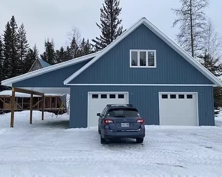 A blue two-storey detached garage with a carport and gable dormer, in a snowy wooded setting