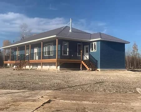 A blue board-and-batten single-storey family home with a full covered front porch, cedar posts and a walkout basement, on a rural lot